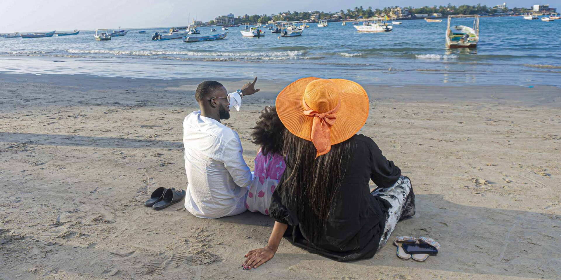 a man and a woman sit on the beach looking at boats in the water