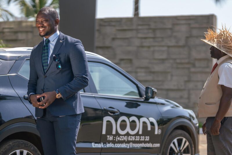 un homme debout à côté d' une voiture noire avec le logo noom